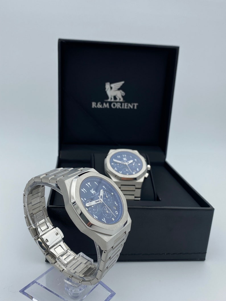 K&ouml;nnte beinhalten: Eine Edelstahl-Armbanduhr mit blauem Zifferblatt und arabischen Ziffern wird neben ihrer Box pr&auml;sentiert. Die Uhr hat ein passendes Edelstahlarmband und wird auf einem transparenten St&auml;nder pr&auml;sentiert. Die Box ist schwarz mit dem Markennamen "R&M ORIENT".