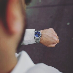 K&ouml;nnte beinhalten: Eine silberne Armbanduhr mit blauem Zifferblatt wird an einem Handgelenk gezeigt. Die Uhr hat ein Metallarmband und ein rundes Zifferblatt mit detailliertem Design. Die Person tr&auml;gt ein wei&szlig;es Hemd.