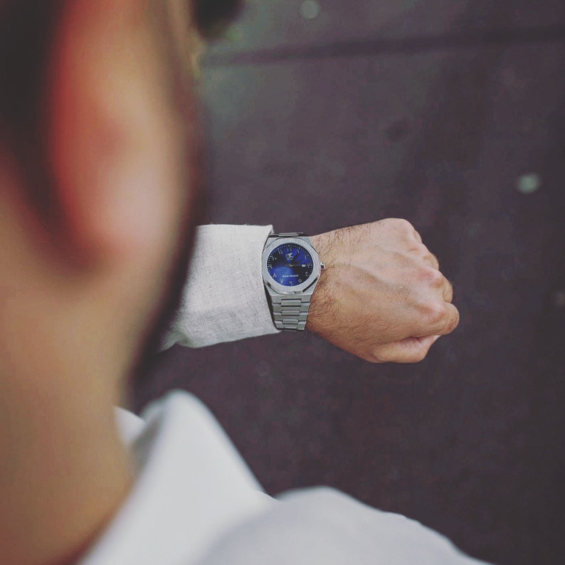 K&ouml;nnte beinhalten: Eine silberne Armbanduhr mit blauem Zifferblatt wird an einem Handgelenk gezeigt. Die Uhr hat ein Metallarmband und ein rundes Zifferblatt mit detailliertem Design. Die Person tr&auml;gt ein wei&szlig;es Hemd.