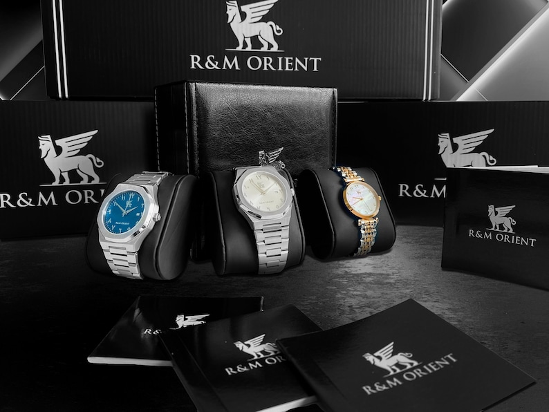 May include: Three wristwatches with silver and gold bands are displayed on black stands. The watches have white, blue, and gold faces. The brand name "R&M ORIENT" is visible on the boxes and booklets in the background.