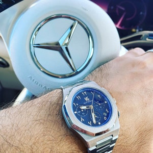 K&ouml;nnte beinhalten: Edelstahl-Armbanduhr mit blauem Zifferblatt und arabischen Ziffern. Die Uhr mit silbernem Armband befindet sich am Handgelenk einer Person. Im Hintergrund ein wei&szlig;es Lenkrad mit silbernem Emblem.