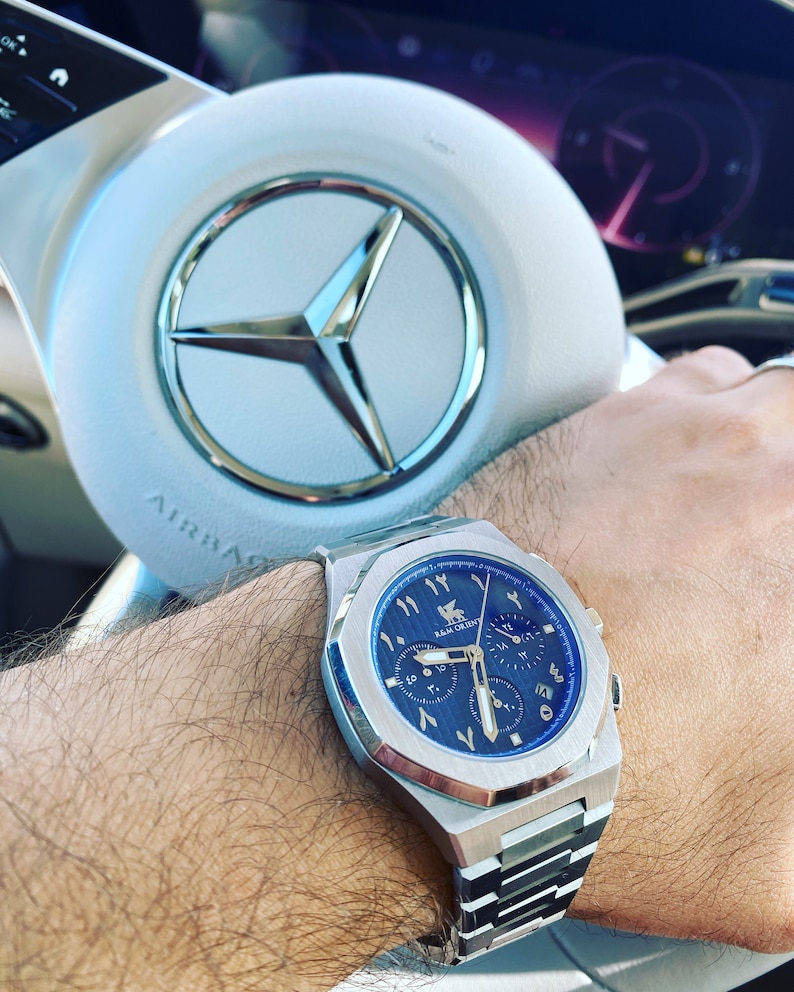 K&ouml;nnte beinhalten: Edelstahl-Armbanduhr mit blauem Zifferblatt und arabischen Ziffern. Die Uhr mit silbernem Armband befindet sich am Handgelenk einer Person. Im Hintergrund ein wei&szlig;es Lenkrad mit silbernem Emblem.