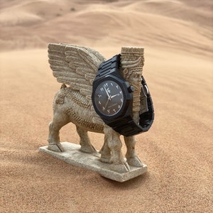 May include: A black wristwatch with Arabic numerals is displayed on a stone sculpture of a winged bull. The watch has a black band and face. The sculpture is set in a desert landscape with sand dunes in the background.