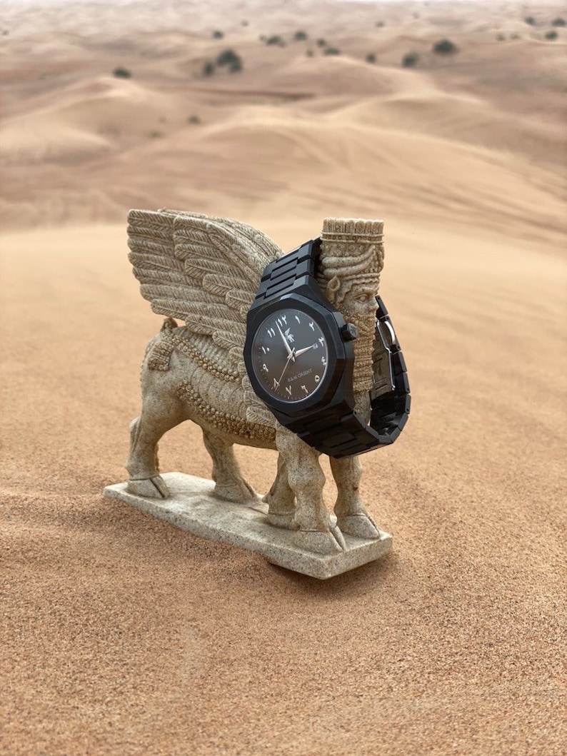 May include: A black wristwatch with Arabic numerals is displayed on a stone sculpture of a winged bull. The watch has a black band and face. The sculpture is set in a desert landscape with sand dunes in the background.