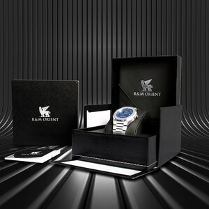 May include: A silver wristwatch with a blue face is displayed in an open black box. The box is embossed with the text "R&M ORIENT" and a winged lion emblem. A closed box with the same logo is to the left, and a booklet is in front.