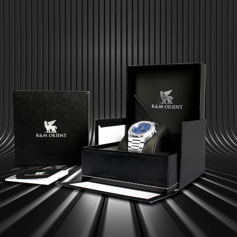 May include: A silver wristwatch with a blue face is displayed in an open black box. The box is embossed with the text "R&M ORIENT" and a winged lion emblem. A closed box with the same logo is to the left, and a booklet is in front.