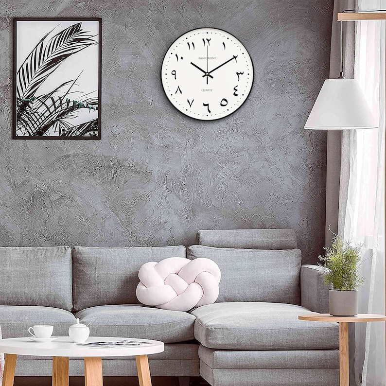 Pu&ograve; includere: Un orologio da parete bianco con numeri arabi neri e una cornice nera. L'orologio &egrave; appeso a una parete grigia sopra un divano grigio con un cuscino rosa.