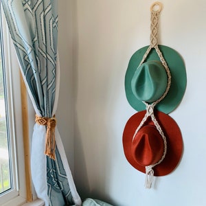 May include: A macrame hat rack with two hats hanging on it. The top hat is green and the bottom hat is red. The rack is made of natural fiber rope and has a wooden ring at the top.