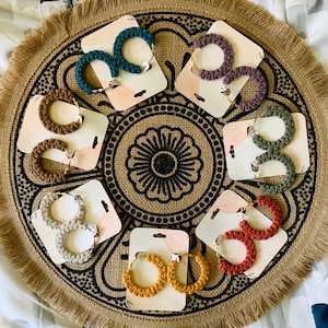 May include: Assortment of eight pairs of crocheted hoop earrings in various colors, including brown, green, purple, and yellow. The earrings are displayed on a circular, brown, textured surface with a black and white mandala design.
