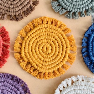 May include: Set of six colorful round crocheted coasters with fringe. The coasters are in shades of brown, blue, yellow, red, white, and purple.