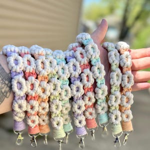 May include: A collection of macrame wristlets in a variety of pastel colors, including pink, orange, yellow, green, and purple. Each wristlet features a flower design and a silver-colored clasp.