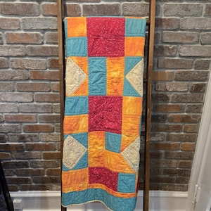 Puede incluir: Un edred&oacute;n de retazos colorido con un patr&oacute;n de estrella, exhibido en una escalera de madera. El edred&oacute;n presenta cuadrados de tela roja, naranja y verde azulado, con detalles de encaje. El edred&oacute;n mide aproximadamente 152 cm de largo.