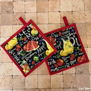 May include: Two square pot holders with a black background and a red border. The pot holders feature illustrations of fruits like watermelon, lemons, and strawberries, along with text that reads "Green Limes" and "Raspberry Apple."