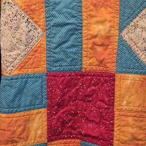 Puede incluir: Primer plano de un edred&oacute;n de retazos con una vibrante mezcla de colores y patrones. El edred&oacute;n presenta cuadrados y rect&aacute;ngulos en tonos de verde azulado, naranja y rojo. Algunos cuadrados tienen un patr&oacute;n de diamantes, otros un dise&ntilde;o moteado.