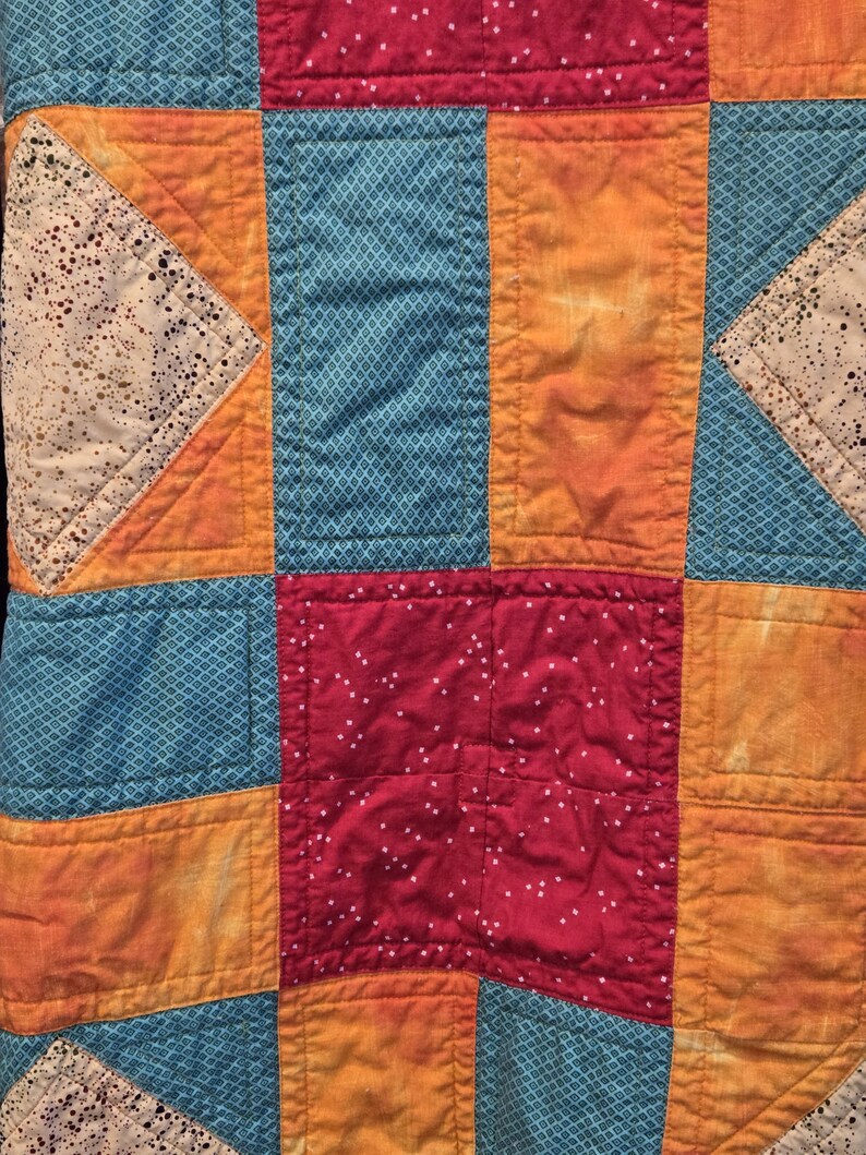 Puede incluir: Primer plano de un edred&oacute;n de retazos con una vibrante mezcla de colores y patrones. El edred&oacute;n presenta cuadrados y rect&aacute;ngulos en tonos de verde azulado, naranja y rojo. Algunos cuadrados tienen un patr&oacute;n de diamantes, otros un dise&ntilde;o moteado.
