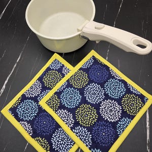 May include: A cream-colored saucepan with a white handle and two navy blue and yellow floral print pot holders. The pot holders have a yellow border and are square-shaped. The background is a black marble-like surface.