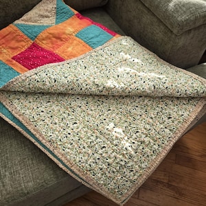 Puede incluir: Un edred&oacute;n hecho a mano con un dise&ntilde;o de patchwork con cuadrados de telas naranja, turquesa, rosa y beige. La parte inferior del edred&oacute;n tiene un estampado de hojas verdes y blancas. Los bordes est&aacute;n acabados con un ribete beige, doblado sobre un sof&aacute; gris.