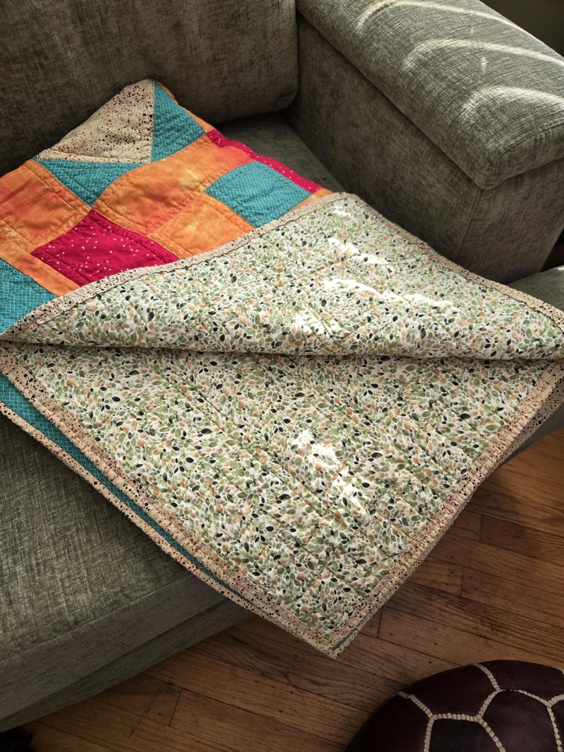 Puede incluir: Un edred&oacute;n hecho a mano con un dise&ntilde;o de patchwork con cuadrados de telas naranja, turquesa, rosa y beige. La parte inferior del edred&oacute;n tiene un estampado de hojas verdes y blancas. Los bordes est&aacute;n acabados con un ribete beige, doblado sobre un sof&aacute; gris.