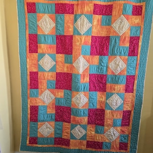 Puede incluir: Un edred&oacute;n colorido con un patr&oacute;n geom&eacute;trico. Presenta cuadrados de color turquesa, naranja y fucsia, con detalles blancos en forma de diamante. El edred&oacute;n est&aacute; bordeado con tela turquesa y colgado de una barra negra.