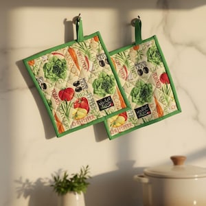 May include: Two square kitchen pot holders with a green border and hanging loop. The cream-colored fabric is patterned with vegetables, including tomatoes, lettuce, carrots, and olives. The text "Market Fresh" is also visible.