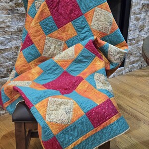 Puede incluir: Un edred&oacute;n de patchwork colorido, drapeado sobre una silla de madera. El edred&oacute;n presenta cuadrados de tela naranja, turquesa y fucsia, con detalles de encaje decorativo. Los bordes del edred&oacute;n est&aacute;n acabados con un borde turquesa.