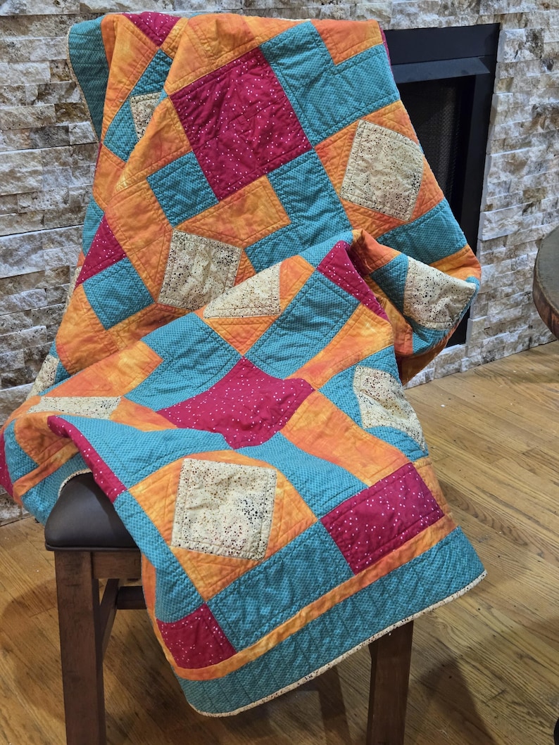 Puede incluir: Un edred&oacute;n de patchwork colorido, drapeado sobre una silla de madera. El edred&oacute;n presenta cuadrados de tela naranja, turquesa y fucsia, con detalles de encaje decorativo. Los bordes del edred&oacute;n est&aacute;n acabados con un borde turquesa.