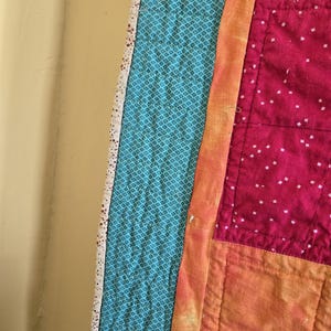 Puede incluir: Primer plano de un edred&oacute;n hecho a mano con un dise&ntilde;o de patchwork. El edred&oacute;n presenta tela turquesa con un patr&oacute;n de diamantes, una franja naranja vibrante y secciones de tela rosa con puntos blancos. Los bordes est&aacute;n acabados con un borde blanco.