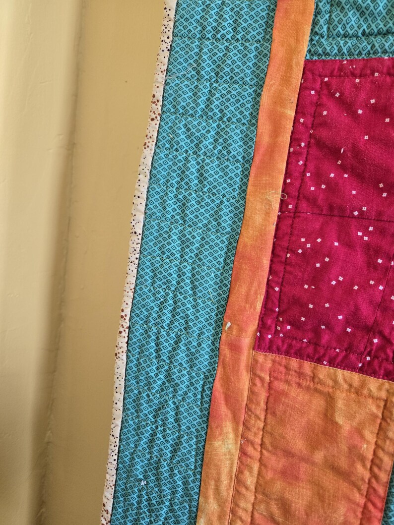 Puede incluir: Primer plano de un edred&oacute;n hecho a mano con un dise&ntilde;o de patchwork. El edred&oacute;n presenta tela turquesa con un patr&oacute;n de diamantes, una franja naranja vibrante y secciones de tela rosa con puntos blancos. Los bordes est&aacute;n acabados con un borde blanco.