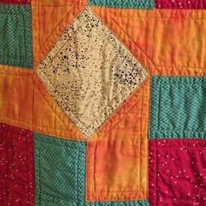 Puede incluir: Primer plano de un edred&oacute;n de retazos con un patr&oacute;n de diamantes en el centro. El edred&oacute;n presenta cuadrados de tela naranja, verde azulado y roja. El diamante central es beige con motas negras. El edred&oacute;n est&aacute; cosido con un patr&oacute;n acolchado.