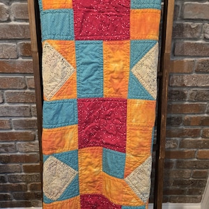Puede incluir: Un edred&oacute;n de retazos colorido con un dise&ntilde;o geom&eacute;trico. El edred&oacute;n presenta cuadrados de tela roja, naranja, turquesa y amarilla, algunos con lunares blancos. Tri&aacute;ngulos de encaje color crema a&ntilde;aden detalles. El edred&oacute;n est&aacute; colgado sobre una escalera de madera.