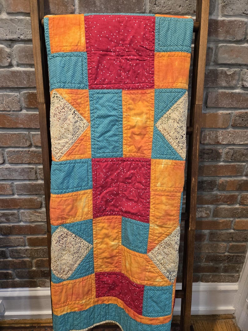 Puede incluir: Un edred&oacute;n de retazos colorido con un dise&ntilde;o geom&eacute;trico. El edred&oacute;n presenta cuadrados de tela roja, naranja, turquesa y amarilla, algunos con lunares blancos. Tri&aacute;ngulos de encaje color crema a&ntilde;aden detalles. El edred&oacute;n est&aacute; colgado sobre una escalera de madera.