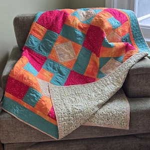 Puede incluir: Una colorida colcha de retazos sobre un sill&oacute;n gris. La colcha presenta cuadrados de tela rosa, naranja y turquesa, con un reverso crema. Los bordes de la colcha est&aacute;n ribeteados con un borde turquesa.