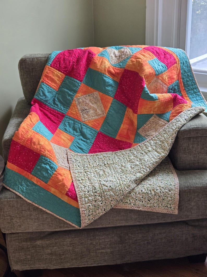 Puede incluir: Una colorida colcha de retazos sobre un sill&oacute;n gris. La colcha presenta cuadrados de tela rosa, naranja y turquesa, con un reverso crema. Los bordes de la colcha est&aacute;n ribeteados con un borde turquesa.