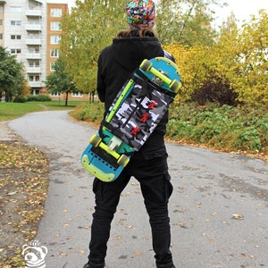 May include: A person wearing a black jacket and black trousers is carrying a skateboard on their back. The skateboard has a camouflage pattern and a blue and yellow design. The person is wearing a colourful hat.