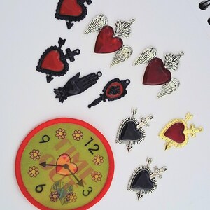 May include: A collection of charms and a patch with a clock design. Charms include red hearts with black accents, winged hearts, and black hearts pierced with arrows. The clock patch is green with a red border and features a heart and butterfly.
