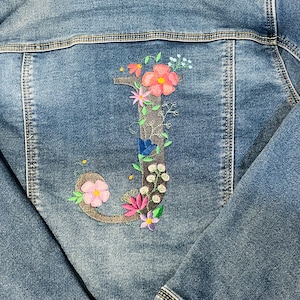 May include: A denim jacket with an embroidered floral design. The back of the jacket features a large, embroidered letter "J" adorned with pink, orange, and blue flowers, and green leaves. The jacket is a medium wash blue.