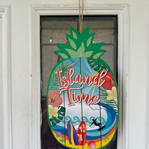 May include: A pineapple-shaped door hanger with the text "Island Time" in red and white lettering. The hanger features a blue and green tropical scene with surfboards, waves, and palm trees.