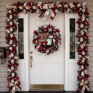 Puede incluir: Una puerta de entrada blanca decorada con una guirnalda y corona navideña con cintas de cuadros rojos, negros y blancos, detalles de arpillera y adornos rojos. La corona incluye un letrero que dice There's Snow place like HOME con un muñeco de nieve.