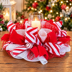Op de afbeelding: Een kersttafelstuk met een doorzichtige glazen cilinder met een witte stompkaars, omringd door een decoratieve krans van rood-wit gestreepte stoffen linten, effen rode fluwelen linten en wit gaasnetwerk.