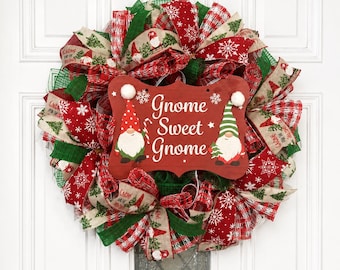 Puede incluir: Una corona navideña con cintas de malla roja, verde y beige con patrones de copos de nieve y gnomos. Un letrero central rojo con borde festoneado muestra el texto Gnome Sweet Gnome, flanqueado por dos gnomos ilustrados con ropa festiva.