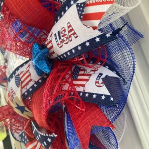 Pu&ograve; includere: Una ghirlanda patriottica rosso, bianco e blu con una stella blu e la parola "USA" su un nastro. La ghirlanda &egrave; fatta di rete e ha un fiocco di juta.