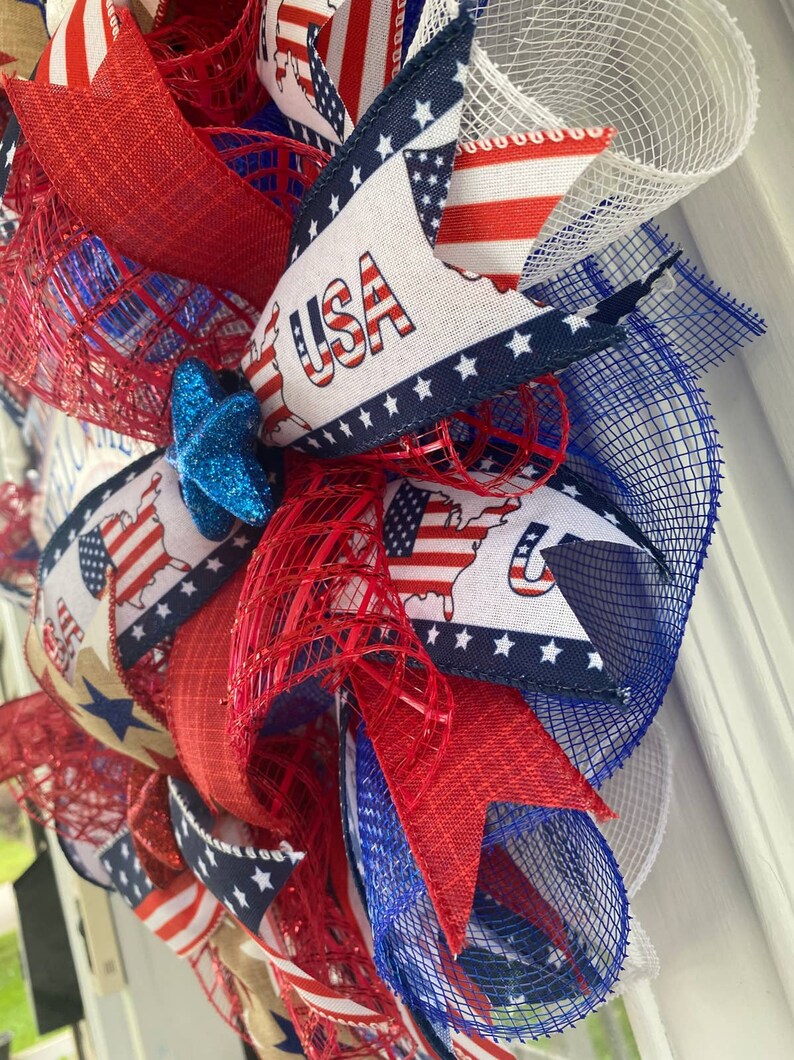 Pu&ograve; includere: Una ghirlanda patriottica rosso, bianco e blu con una stella blu e la parola "USA" su un nastro. La ghirlanda &egrave; fatta di rete e ha un fiocco di juta.