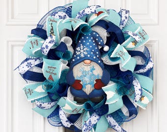 Puede incluir: Una corona decorativa con una figura central de gnomo que lleva un sombrero azul con lunares blancos y un patrón de copos de nieve. La corona está hecha de capas de cintas de malla azul, azul claro y blanca con estampados de copos y texto.