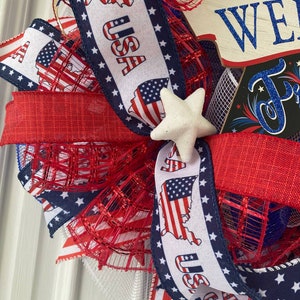 Pu&ograve; includere: Una ghirlanda patriottica rosso, bianco e blu con un cartello che dice "America We Love You" e una stella bianca. La ghirlanda &egrave; realizzata con rete e nastro rosso, bianco e blu con la scritta "USA" stampata su di essa.