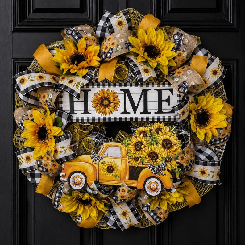 Op de afbeelding: Een decoratieve krans met gele zonnebloemen, zwart-wit geblokte linten en een centraal bord met het woord HOME, waarbij de letter O is vervangen door een zonnebloem. Een gele vrachtwagen met zonnebloemen is aan de onderkant bevestigd.