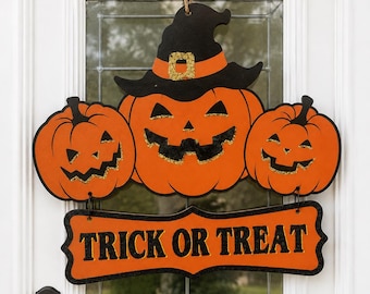 May include: A Halloween door sign featuring three orange jack-o'-lanterns, with the centre pumpkin wearing a black witch hat with an orange band. Below the pumpkins, an orange sign with black glittery edges displays the text TRICK OR TREAT.