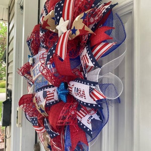 Pu&ograve; includere: Una ghirlanda patriottica rosso, bianco e blu con un cartello che dice "Bless America, Welcome Home, Land of the Free". La ghirlanda &egrave; decorata con stelle, strisce e un fiocco blu.