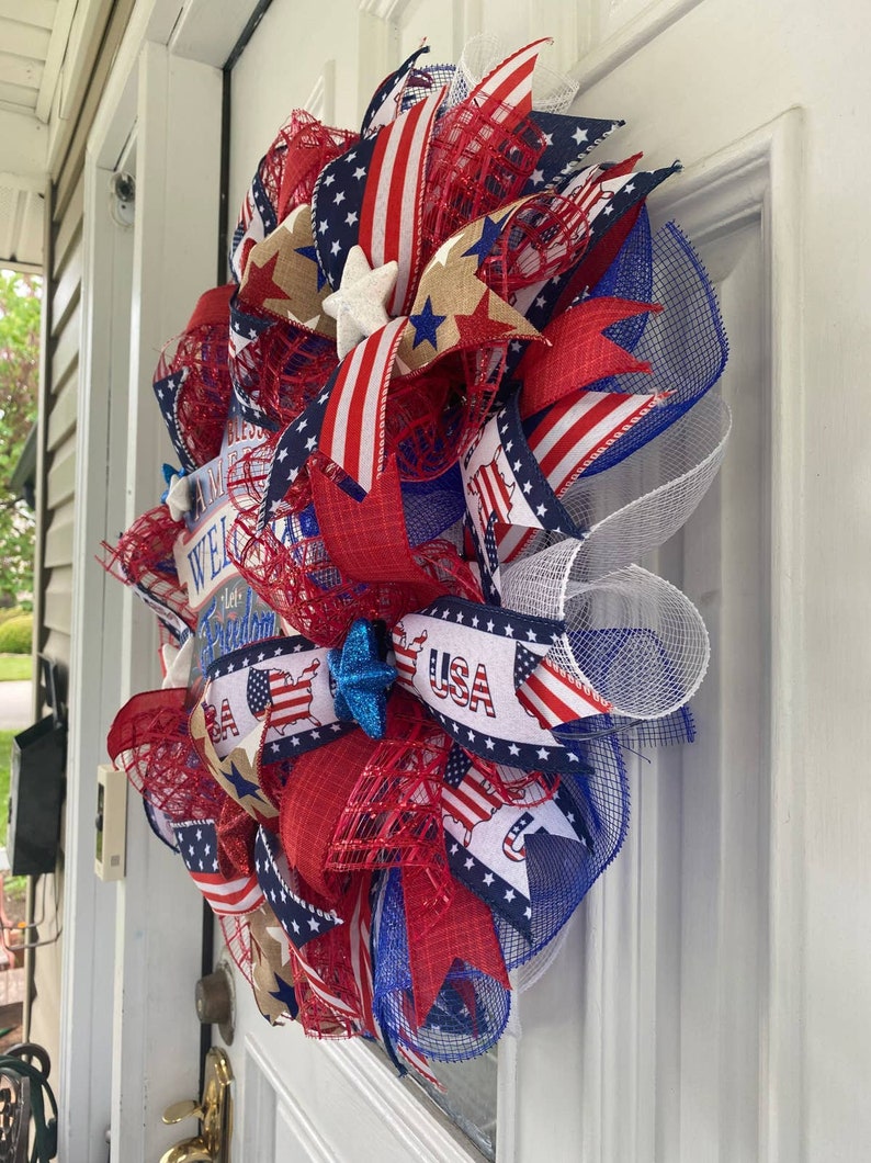 Pu&ograve; includere: Una ghirlanda patriottica rosso, bianco e blu con un cartello che dice "Bless America, Welcome Home, Land of the Free". La ghirlanda &egrave; decorata con stelle, strisce e un fiocco blu.