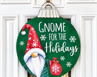 Puede incluir: Un letrero redondo de madera verde con forma de adorno navideño y tapa de metal plateado. Muestra un gnomo con gorro rojo de copos blancos, barba blanca y un saco rojo. El texto Gnome for the Holidays aparece escrito en color blanco.