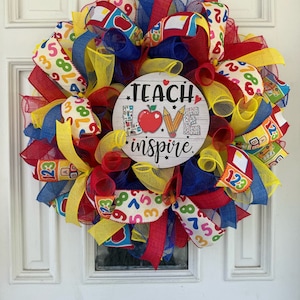 Op de afbeelding: Een kleurrijke leraar krans met rode, gele en blauwe linten en gaas. De krans heeft een houten bord in het midden met de tekst "Teach Love Inspire".